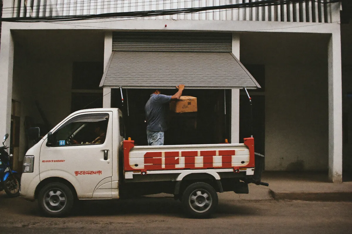 Two men lifting a carton from the tailgate of a small delivery truck backed up to a shop shutter on a street.