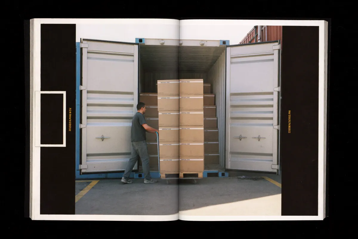 Cartons on a hand pallet truck being loaded into the open rear doors of a shipping container at a dock.
