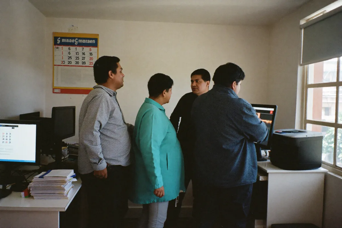 Three colleagues in a small office looking at a monitor together, box files and a printer on the desk.