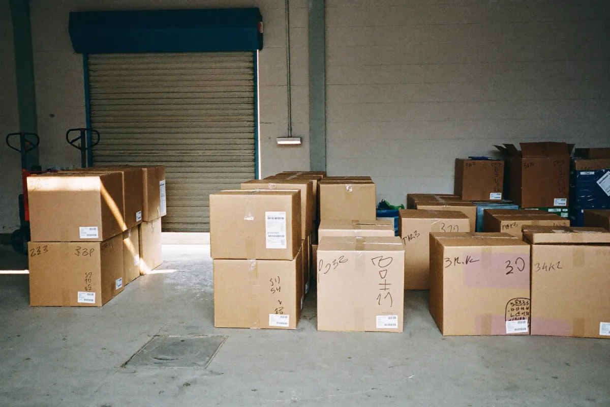 Cardboard cartons stacked in lots on the floor of a goods-consolidation warehouse, roller shutter open at the back.